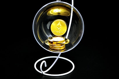 A stack of gold-colored Ethereum coins placed inside a transparent spherical container held by a white, spiral stand on a black background.