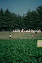 A lush green park with a large, well-maintained lawn where several children are running and playing. In the background, a row of small white tents is visible, and tall evergreen trees create a natural wall. The foreground features a dense patch of greenery. The image conveys a sense of outdoor recreation and freedom.