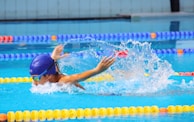 A swimmer wearing a blue swim cap and goggles is performing a butterfly stroke in a pool with clear blue water. Splashes of water surround the swimmer, and lane dividers in vibrant colors such as yellow, blue, and orange are visible.