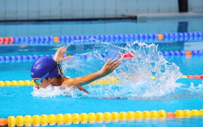 A swimmer wearing a blue swim cap and goggles is performing a butterfly stroke in a pool with clear blue water. Splashes of water surround the swimmer, and lane dividers in vibrant colors such as yellow, blue, and orange are visible.