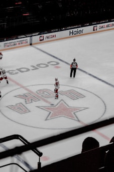 An ice hockey match is underway in an arena with players in jerseys and an official in black and white stripes visible. The center of the rink features a large star emblem with Russian text around it. Advertisements in Cyrillic script are displayed on the boards surrounding the rink.