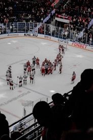 A group of ice hockey players in red and white jerseys gather on the ice, surrounded by referees. The stadium is filled with spectators, some standing, watching from behind protective glass. Advertisements and team logos adorn the rink and surrounding walls. Bright lighting illuminates the scene from above.