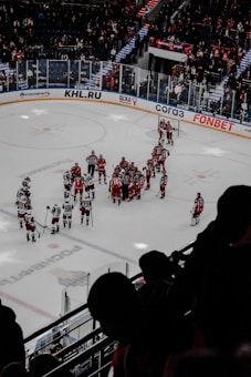 A group of ice hockey players in red and white jerseys gather on the ice, surrounded by referees. The stadium is filled with spectators, some standing, watching from behind protective glass. Advertisements and team logos adorn the rink and surrounding walls. Bright lighting illuminates the scene from above.