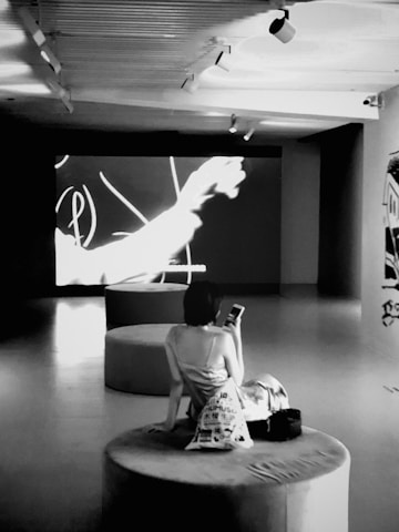 A person is sitting on a circular cushioned seat within a dimly lit room, facing a large screen showing abstract shapes and lines. The person holds a phone in one hand and has a bag placed beside them. The room has a modern industrial feel with overhead spotlights and clean lines.