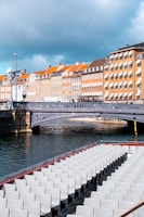 A scenic view of a canal lined with classic European-style buildings featuring distinct orange rooftops and numerous windows. A bridge crosses the canal, and in the forefront, an empty tourist boat with multiple rows of white seats is visible on the water.