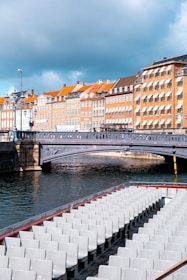 A scenic view of a canal lined with classic European-style buildings featuring distinct orange rooftops and numerous windows. A bridge crosses the canal, and in the forefront, an empty tourist boat with multiple rows of white seats is visible on the water.