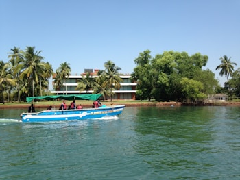 A motorboat with several passengers wearing life vests travels across a calm body of water. In the background, a building with a modern design is partially obscured by palm trees and other lush green vegetation. The sky is clear and blue.
