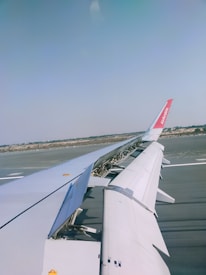 The photo captures an airplane wing with extended flaps and a view of the tarmac below. The landscape beyond the runway is visible, along with clear blue skies.