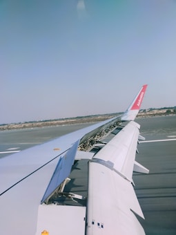 The photo captures an airplane wing with extended flaps and a view of the tarmac below. The landscape beyond the runway is visible, along with clear blue skies.