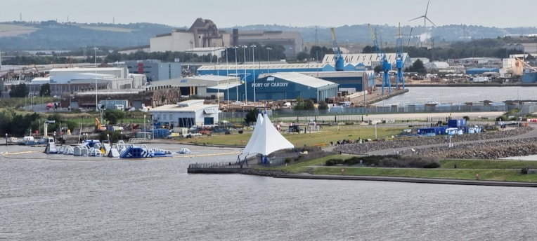 A waterfront industrial area featuring large buildings and cranes, with a prominent marquee tent near the water. The setup includes various smaller structures and an inflatable obstacle course on the water, all situated under a partly cloudy sky. In the background, wind turbines are visible on the rolling hills.