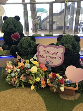 A decorative display featuring topiary figures shaped like pandas, surrounded by colorful flowers. A sign reads 'Happy Mother's Day,' accompanied by a small heart decoration. The scene is set indoors with visible windows and soft lighting.