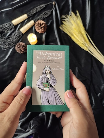 Hands are holding a green box labeled 'The Alchemical Tarot: Renewed 5th Edition' featuring an illustration of a figure in robes. The background includes various objects such as pine cones, a lit candle, wheat stalks, and decorative tassels placed on a black fabric.