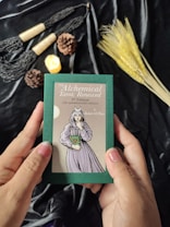 Hands are holding a green box labeled 'The Alchemical Tarot: Renewed 5th Edition' featuring an illustration of a figure in robes. The background includes various objects such as pine cones, a lit candle, wheat stalks, and decorative tassels placed on a black fabric.