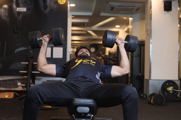 A person is lying on a bench in a gym environment, lifting heavy dumbbells with both hands. The individual is wearing a black workout shirt with a yellow print and appears focused on their exercise. The background includes gym equipment and mirrors, creating an active and motivational setting.