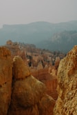 An expansive view of rugged, orange and tan-colored rock formations creating a canyon landscape. Eroded spires, known as hoodoos, are visible in the midground, and the distant backdrop features densely forested plateaus under a hazy sky.