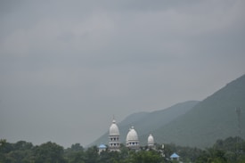 A serene landscape featuring a temple with three white domes partially visible amidst lush greenery. The backdrop includes misty hills under a cloudy, gray sky, creating a tranquil and calm atmosphere.