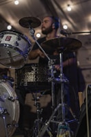 A person is playing the drums with focused expression under ambient lighting. The drum set includes a bass drum, snare drum, and cymbals. The scene appears to be indoors with soft, warm lighting providing a cozy atmosphere.