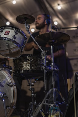 A person is playing the drums with focused expression under ambient lighting. The drum set includes a bass drum, snare drum, and cymbals. The scene appears to be indoors with soft, warm lighting providing a cozy atmosphere.