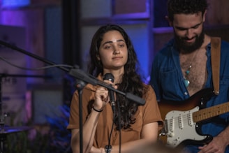 A woman holding a microphone appears to be engaged in a performance or speech. A man in the background with a guitar seems to be part of a musical setup. The setting has a cozy, indoor ambiance with a mix of blue and purple lighting.