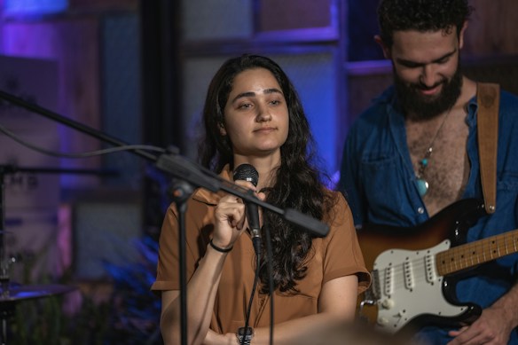 A woman holding a microphone appears to be engaged in a performance or speech. A man in the background with a guitar seems to be part of a musical setup. The setting has a cozy, indoor ambiance with a mix of blue and purple lighting.