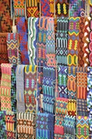 A vibrant array of intricately woven textile patterns featuring geometric designs. The textiles display a rich variety of colors and intricate details, arranged in rows and showcasing a range of traditional and cultural motifs.