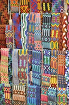 A vibrant array of intricately woven textile patterns featuring geometric designs. The textiles display a rich variety of colors and intricate details, arranged in rows and showcasing a range of traditional and cultural motifs.