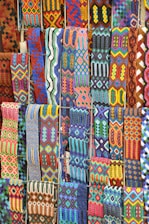 A vibrant array of intricately woven textile patterns featuring geometric designs. The textiles display a rich variety of colors and intricate details, arranged in rows and showcasing a range of traditional and cultural motifs.