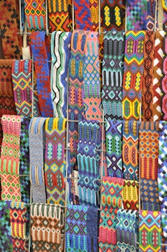 A vibrant array of intricately woven textile patterns featuring geometric designs. The textiles display a rich variety of colors and intricate details, arranged in rows and showcasing a range of traditional and cultural motifs.