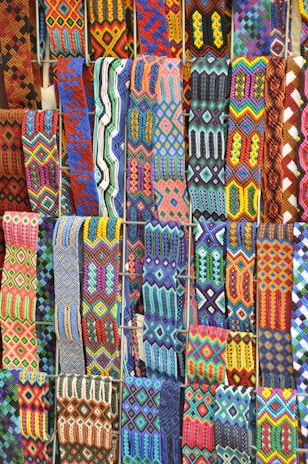 A vibrant array of intricately woven textile patterns featuring geometric designs. The textiles display a rich variety of colors and intricate details, arranged in rows and showcasing a range of traditional and cultural motifs.