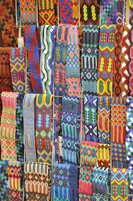 A vibrant array of intricately woven textile patterns featuring geometric designs. The textiles display a rich variety of colors and intricate details, arranged in rows and showcasing a range of traditional and cultural motifs.
