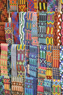 A vibrant array of intricately woven textile patterns featuring geometric designs. The textiles display a rich variety of colors and intricate details, arranged in rows and showcasing a range of traditional and cultural motifs.