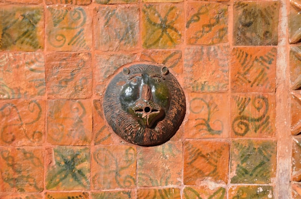 A ceramic artwork depicting a lion's head is mounted on a richly patterned terracotta tile wall. The tiles feature intricate green and orange motifs, adding to the rustic and artistic feel of the piece.