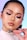 A person with expertly applied makeup, featuring pink and shimmering eyeshadow, defined eyeliner, and glossy lips. They hold a pink-toned cosmetic product labeled 'Rare Beauty,'. The background is softly blurred with hues of blue and pink.