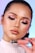 A person with expertly applied makeup, featuring pink and shimmering eyeshadow, defined eyeliner, and glossy lips. They hold a pink-toned cosmetic product labeled 'Rare Beauty,'. The background is softly blurred with hues of blue and pink.
