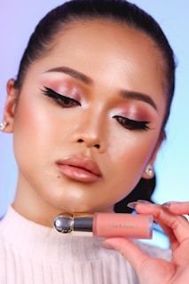 A person with expertly applied makeup, featuring pink and shimmering eyeshadow, defined eyeliner, and glossy lips. They hold a pink-toned cosmetic product labeled 'Rare Beauty,'. The background is softly blurred with hues of blue and pink.