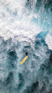 A surfer navigates a powerful wave with a bright yellow surfboard. The ocean is a mix of swirling white foam and deep blue waters, capturing the dynamic motion and energy of the sea.