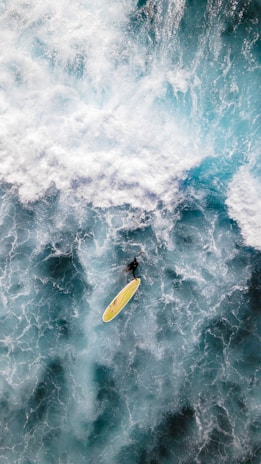 A surfer navigates a powerful wave with a bright yellow surfboard. The ocean is a mix of swirling white foam and deep blue waters, capturing the dynamic motion and energy of the sea.