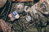 A collection of military items placed on camouflage fabric includes medals, patches, bullets, a tactical cap with a 'Don't Tread on Me' patch, and a brown holster.