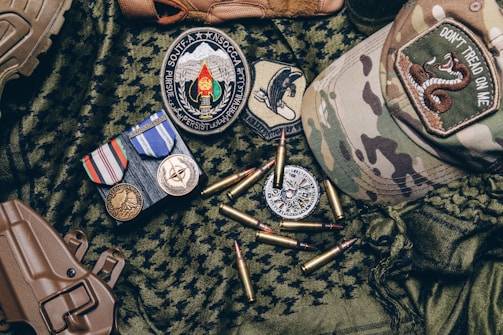 A collection of military items placed on camouflage fabric includes medals, patches, bullets, a tactical cap with a 'Don't Tread on Me' patch, and a brown holster.