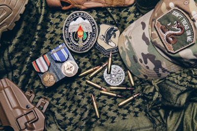 A collection of military items placed on camouflage fabric includes medals, patches, bullets, a tactical cap with a 'Don't Tread on Me' patch, and a brown holster.