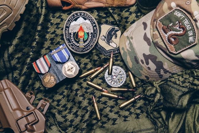A collection of military items placed on camouflage fabric includes medals, patches, bullets, a tactical cap with a 'Don't Tread on Me' patch, and a brown holster.