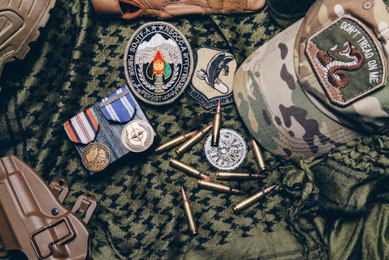 A collection of military items placed on camouflage fabric includes medals, patches, bullets, a tactical cap with a 'Don't Tread on Me' patch, and a brown holster.