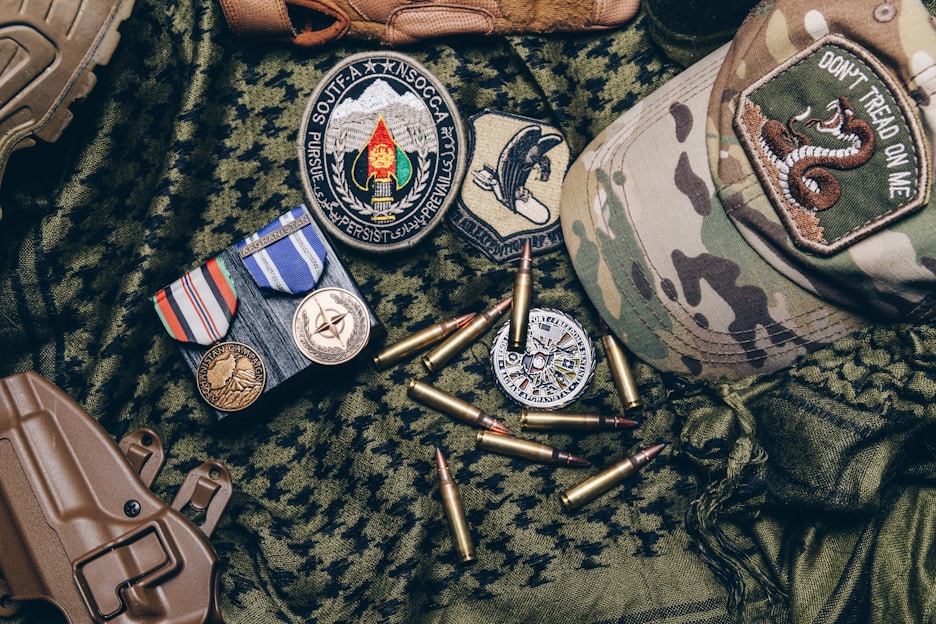 A collection of military items placed on camouflage fabric includes medals, patches, bullets, a tactical cap with a 'Don't Tread on Me' patch, and a brown holster.