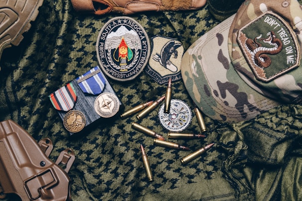 A collection of military items placed on camouflage fabric includes medals, patches, bullets, a tactical cap with a 'Don't Tread on Me' patch, and a brown holster.
