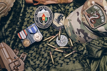 A collection of military items placed on camouflage fabric includes medals, patches, bullets, a tactical cap with a 'Don't Tread on Me' patch, and a brown holster.