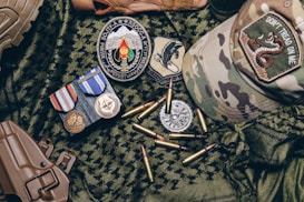 A collection of military items placed on camouflage fabric includes medals, patches, bullets, a tactical cap with a 'Don't Tread on Me' patch, and a brown holster.