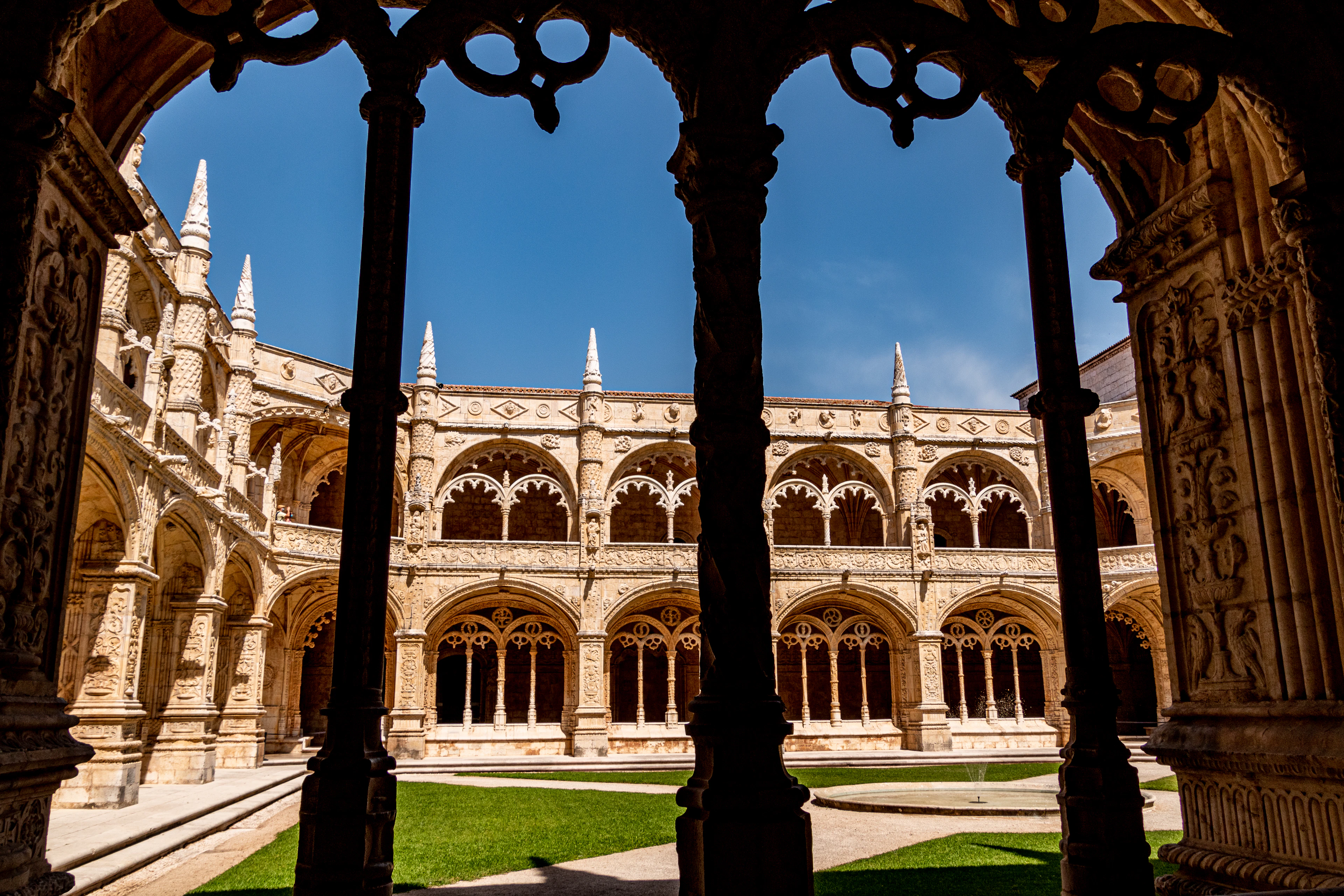 Gothic stone arches of the Jerónimos Monastery cloister framing a sunlit courtyard garden.