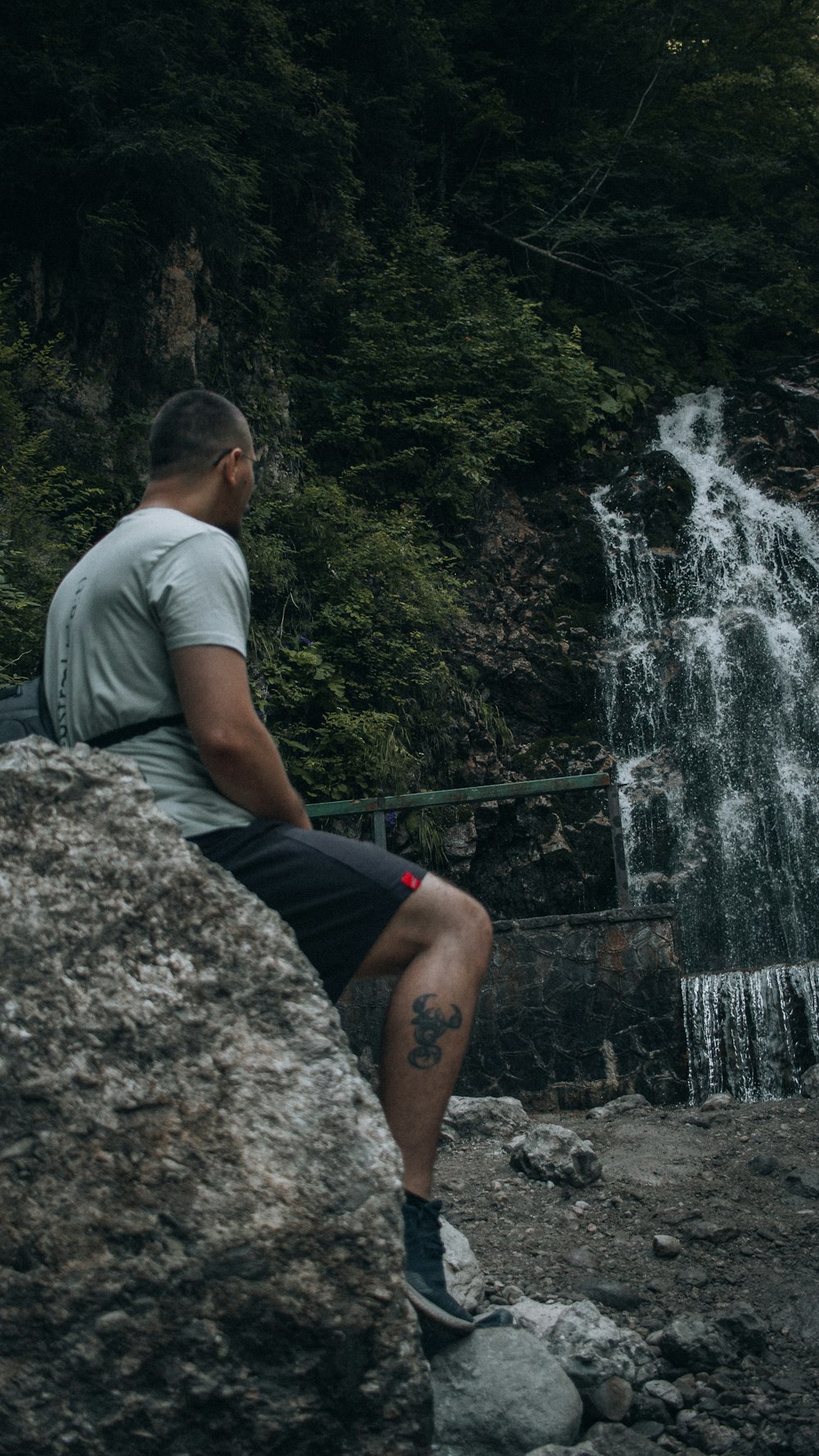 man in gray t-shirt and black shorts standing on rock near waterfalls during daytime