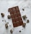 A chocolate bar with a grid pattern is centered on a white marble surface, surrounded by chocolate chunks and crumbs scattered around.