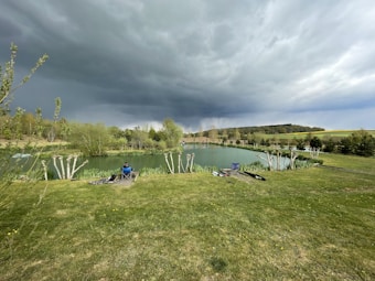 A person sits in a folding chair by a tranquil lake surrounded by lush greenery under a dramatic, cloudy sky. Fishing gear is scattered on the grass, which is bordered by reeds. In the distance, rolling hills and trees add depth to the landscape.